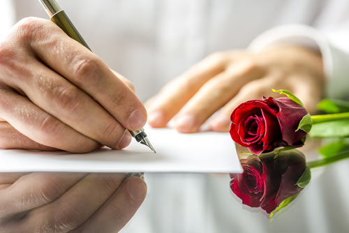 Close-up of a man's hands. He holds a fountain pen in one and is writing a love letter. A red rose is touching an edge of the paper.