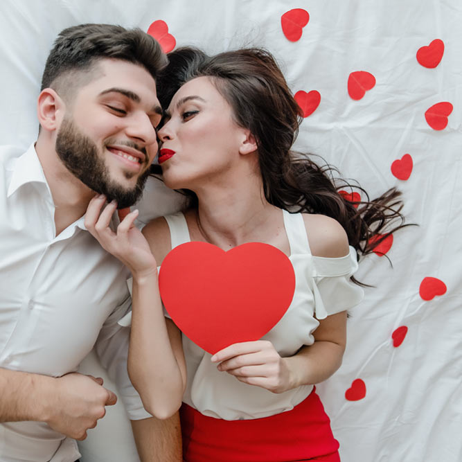 This Romance Jumpstarters image shows a clothed couple laying atop white linen sheets. The woman is holding a large red heart in her left hand and planting a kiss on the cheek of the man who is smiling with delight.