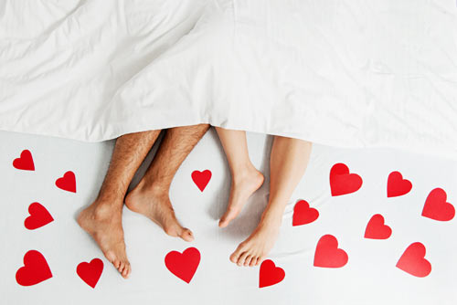 Bare legs of a man and a woman poke out from beneath a bed sheet. Surrounding their feet are red hearts of various sizes.